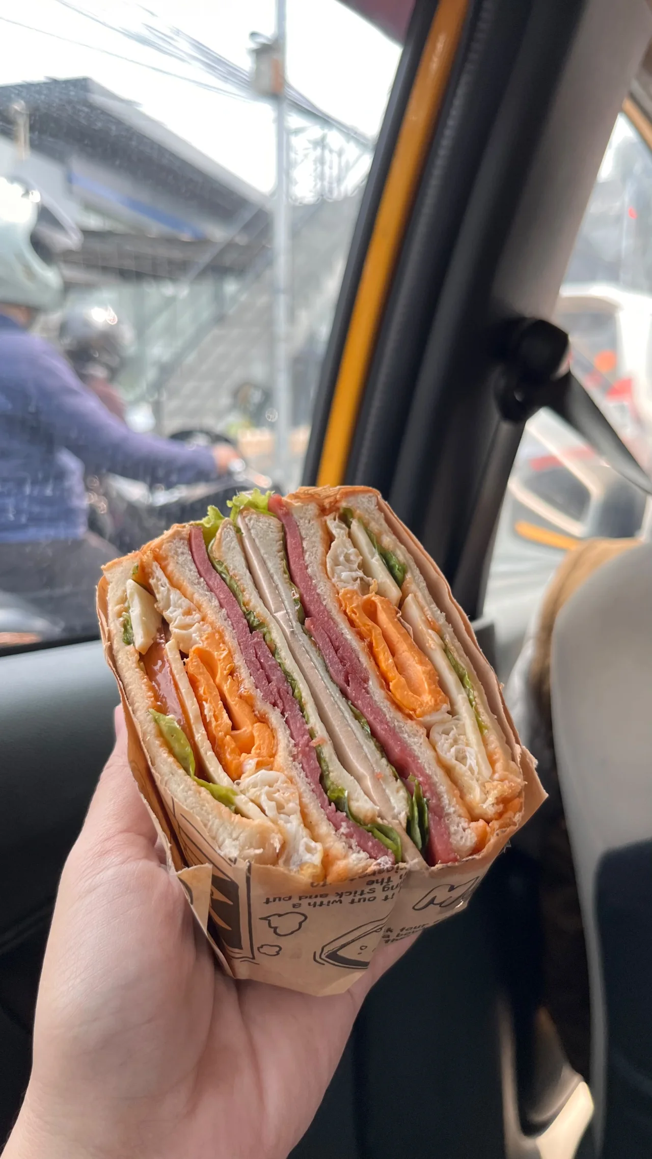 Wanpaku Sandwich berisi lapisan sayur, telur, dan isian lainnya, dipegang di dalam mobil